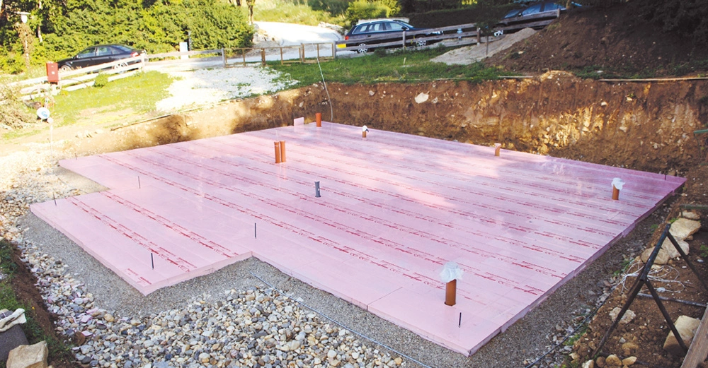 Rosa Dämmplatten liegen auf einem vorbereiteten Bauplatz während des Hausbaus mit Schotterumgebung und geparkten Autos auf einer Schotterstraße im Hintergrund.