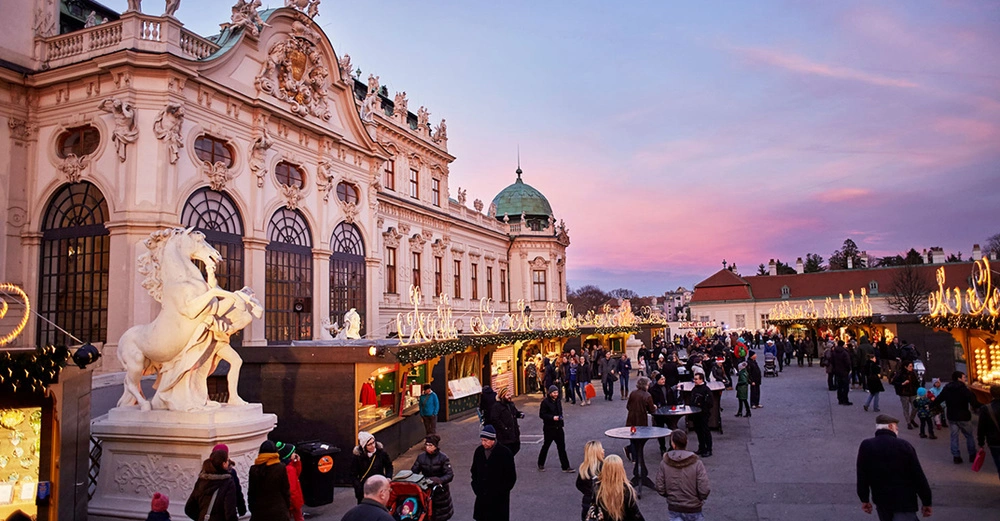 Weihnachtsdorf vor dem Schloss Belvedere in Wien bei violetter Abenddämmerung mit beleuchteten Marktständen.