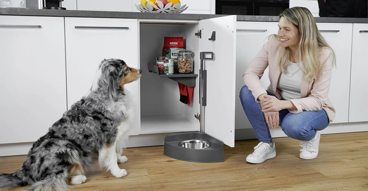 Hund sitzt Küchenboden Frau hockt daneben Küchenschrank geöffnet Hundefutter und Leckerli auf Regalen in Schrank Hundeschale am Boden moderner Küchenhintergrund Obstschale auf Arbeitsplatte.