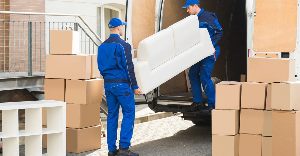 Zwei Männer in blauer Arbeitskleidung laden ein weißes Sofa aus einem Lieferwagen aus umgeben von gestapelten Umzugskartons und einem weißen Regal vor einem Wohngebäude.