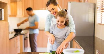 Frau umarmt und hilft Mädchen beim Gemüse schneiden Küche mit Mann der am Herd kocht helle Holzmöbel Kühlschrank links Fenster mit Jalousien rechts