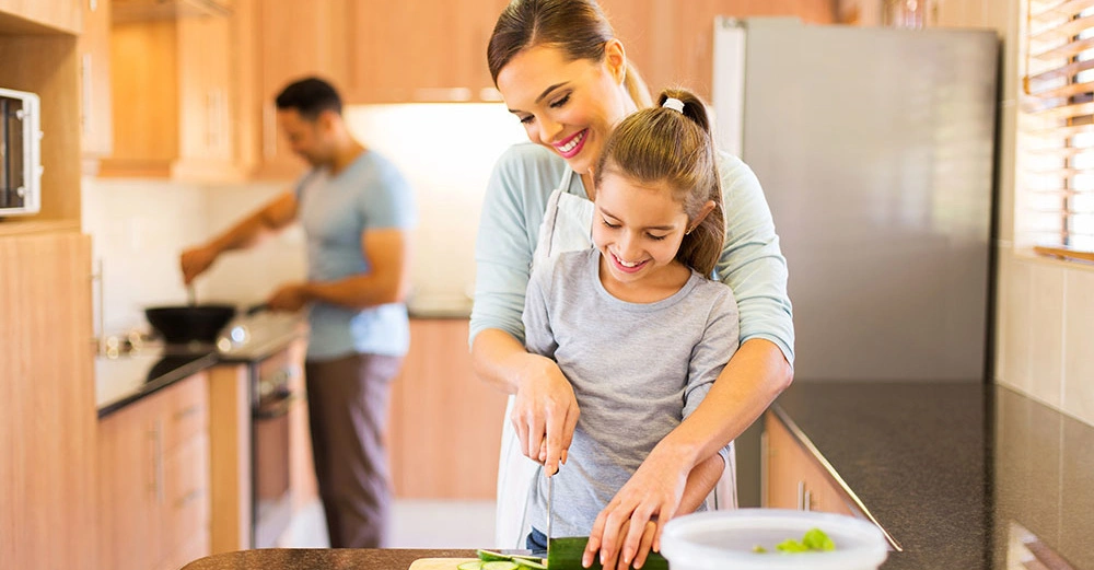 Frau umarmt und hilft Mädchen beim Gemüse schneiden Küche mit Mann der am Herd kocht helle Holzmöbel Kühlschrank links Fenster mit Jalousien rechts