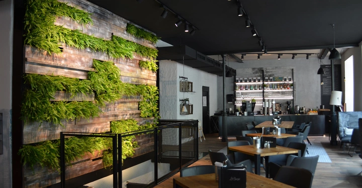 Pflanzenwand bedeckt eine Holzwand in einem modern eingerichteten Café mit Holztischen, schwarzen Stühlen und einer Bar im Hintergrund. Kegelbeleuchtung an schwarzer Decke illuminieren den Raum.