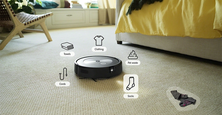 Staubsaugerroboter auf einem hellen Teppich erkennt verschiedene Objekte wie Kleidung Handtücher Haustierabfälle Kabel und Socken in einem hellen Wohnzimmer neben einem gelben Bett.