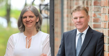 Eine Frau lächelt und trägt eine weiße Bluse im Freien vor einer verschwommenen grünen Landschaft. Ein Mann in einem Anzug lehnt an einer Ziegelwand.