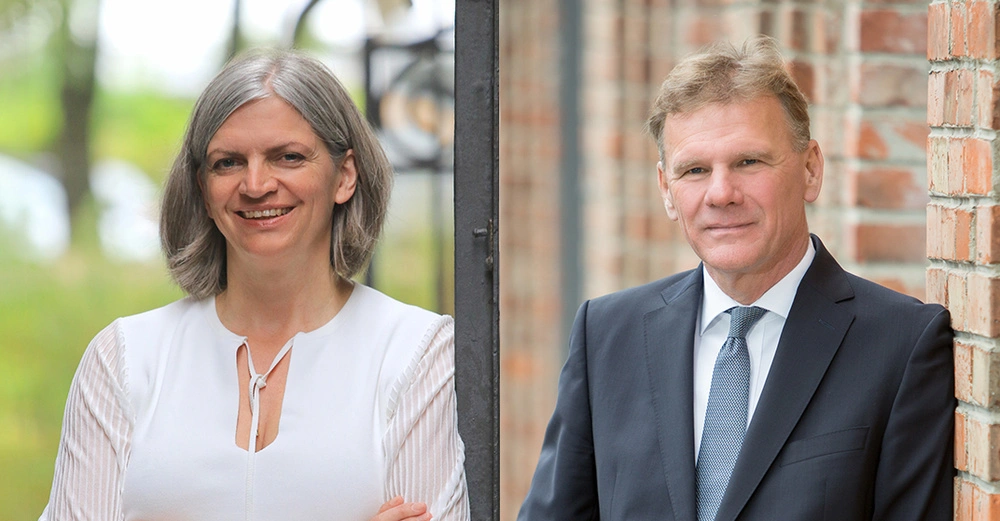 Eine Frau lächelt und trägt eine weiße Bluse im Freien vor einer verschwommenen grünen Landschaft. Ein Mann in einem Anzug lehnt an einer Ziegelwand.