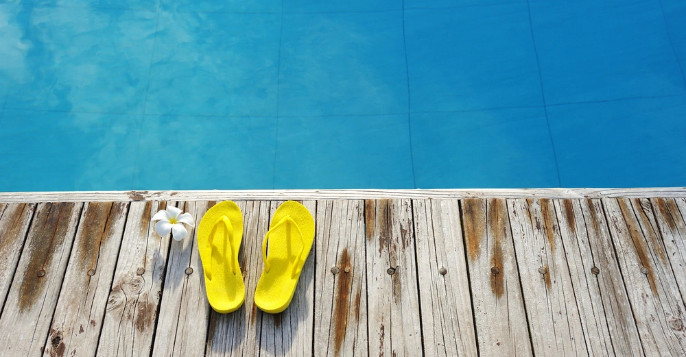 Gelbe Flip-Flops und eine weiße Blüte liegen auf einem hölzernen Steg am Rand eines klaren blauen Pools