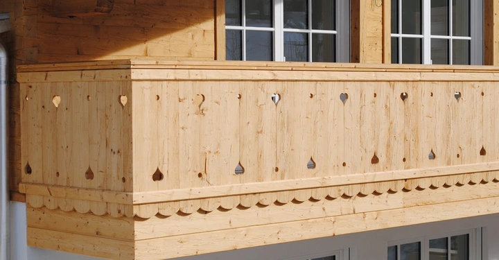 Holzbalkon verziert mit herzförmigen Ausschnitten hängt an einem Gebäude mit weißen Fensterrahmen in einem sonnigen Außenbereich.