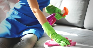 Person trägt grüne Handschuhe und reinigt ein graues Sofa mit einem pinken Tuch und einer Sprühflasche in einem gut beleuchteten Raum.