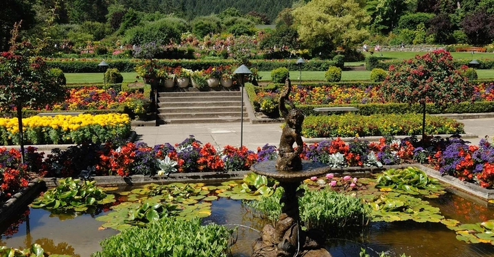 Springbrunnenfigur sprüht Wasser im Teich voller Seerosen in einem gepflegten Blumenpark umgeben von bunten Blumen und üppigem Grün. Im Hintergrund sind Treppen und Waldhügel sichtbar.
