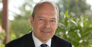 Mann lächelt leicht sitzt in einem Anzug mit Krawatte in einem grünen natürlichen Außenbereich mit Pflanzen im Hintergrund