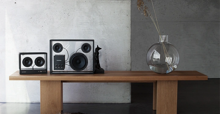 Lautsprecher stehen auf einer modernen Holzbank vor einer Betonwand. Rechts daneben eine Glasvase mit Wasser und Trockenblumen. Ein kleiner Skulptur ist zwischen den Lautsprechern platziert.