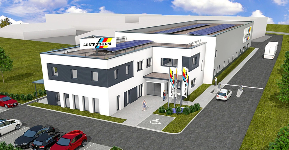 Ein modernes zweistöckiges Bürogebäude mit dem Schriftzug AUSTROTHERM auf einem Schild und Fahnen steht neben einem großen Lagerhaus. Im Vordergrund sind Parkplätze und Menschen zu sehen.