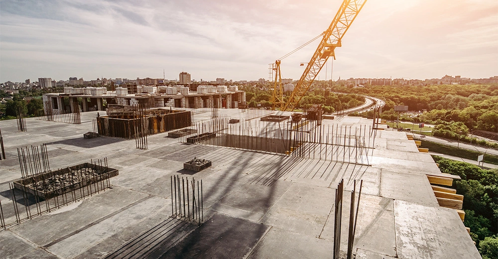 Baustelle mit einem gelben Kran im Einsatz auf einem Gebäude mit Stahldrahtstangen umgeben von Bäumen und einer städtischen Skyline im Hintergrund