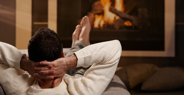 Ein Mann liegt entspannt auf einem Sofa, die Hände hinter dem Kopf verschränkt, und blickt in ein loderndes Kaminfeuer. Kissen liegen auf dem Boden im Hintergrund.