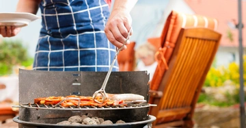 Person in Schürze grillt Fleisch und Wurst mit Zange im Garten mit Holzmöbeln im Hintergrund