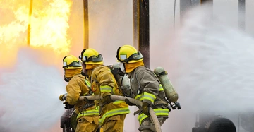Feuerwehrleute in Schutzkleidung und Helmen verwenden Schläuche zum Löschen eines großen Feuers in einer industriellen Umgebung mit viel Rauch und Flammen im Hintergrund.