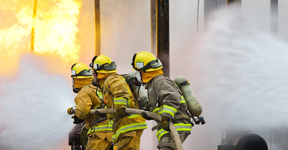 Feuerwehrleute in Schutzkleidung und Helmen verwenden Schläuche zum Löschen eines großen Feuers in einer industriellen Umgebung mit viel Rauch und Flammen im Hintergrund.