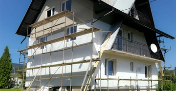 Haus mit Baugerüsten in Renovierung; daneben eine grüne Wiese; sonniger Tag und fahrbare Geräte am Boden.