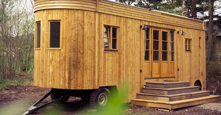 Galerie-Bild 11: Holzanhänger an einem Anhängerhaken stehend umgeben von Bäumen auf unbefestigtem Boden mit Treppe vor der Eingangstür und mehreren Fenstern.