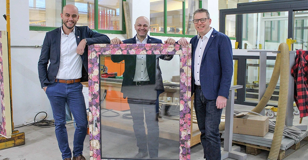 Drei Männer posieren lächelnd in einer Werkstatt und halten gemeinsam einen großen rechteckigen Bilderrahmen mit Blumendesign umrandet vor einer Werkstatt hinter ihnen mit verschiedenen Werkzeugen und Maschinen