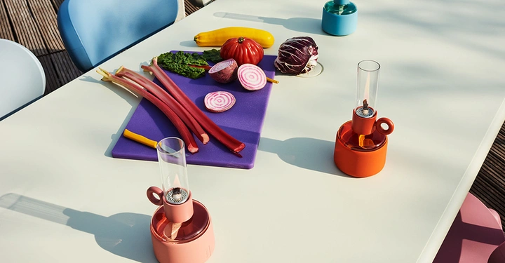 Gemüse liegt auf einem weißen Tisch mit Schneidebrett und kleinen Lampen umgeben von bunten Stühlen im Freien