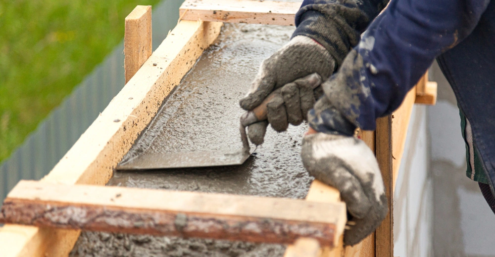 Eine Person verdichtet und nivelliert mit einer Kelle den flüssigen Beton in einer Betonschalung. Die Person trägt helle, vom Beton verschmutzte Arbeitshandschuhe.