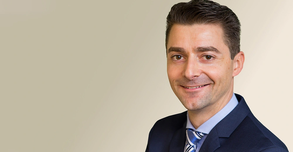 Mann trägt Anzug lächelt leicht befindet sich vor beigem Hintergrund