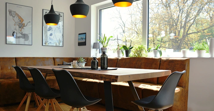 Ein rechteckiger Holztisch steht in einem modernen Essbereich. Schwarze Stühle und Ledersitzbänke drumherum. Drei hängende Lampen beleuchten den Tisch. Fenster und Pflanzen im Hintergrund. An der Wand hängen drei Bilder.