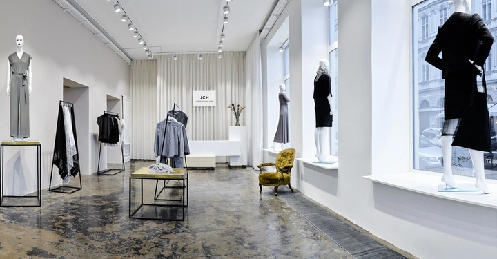 Galerie-Bild 3: Schaufensterpuppen tragen Kleidung in einem minimalistischen Geschäft mit weißen Wänden, hohen Decken und großen Fenstern. Der Boden ist industrieartig und an Wänden hängen weitere Kleidungsstücke. Ein Stuhl steht nahe den Fenstern. Text: JCH.