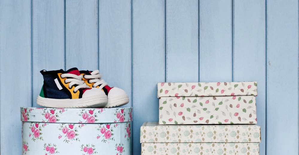 Mehrfarbige Babyturnschuhe stehen auf einer runden Blumenbox daneben drei gestapelte Blumenboxen vor einer hellblauen Holzwand.