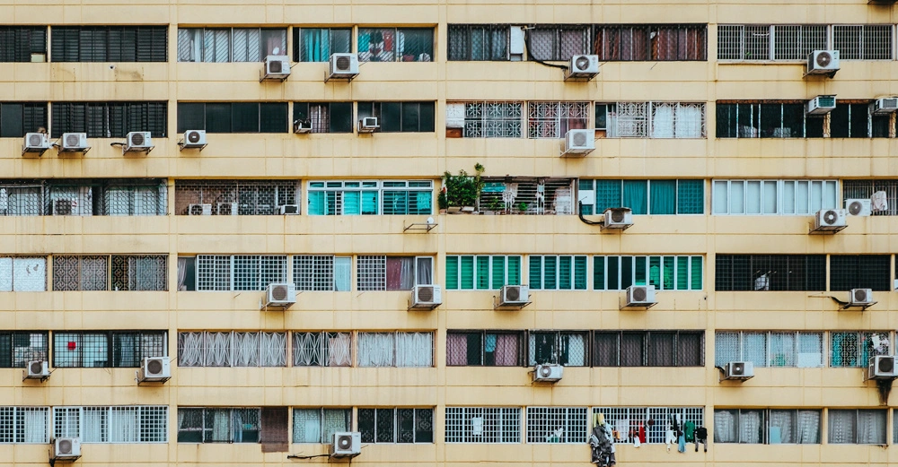 Mehrstöckiges Gebäude mit gelber Fassade und zahlreichen Fenstern Klimaanlagen an fast jedem Fenster Waschleinen mit Kleidung Sichtbar Wohnungen mit unterschiedlichen Vorhängen Stadtumgebung