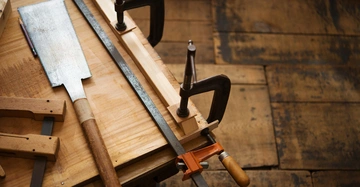 Handsägen und Schraubzwingen liegen auf einer Werkbank in einer Holzbearbeitungswerkstatt. Die Umgebung zeigt einen Holzboden, der dem Raum eine rustikale Atmosphäre verleiht.