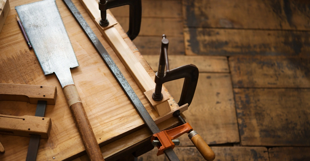 Handsägen und Schraubzwingen liegen auf einer Werkbank in einer Holzbearbeitungswerkstatt. Die Umgebung zeigt einen Holzboden, der dem Raum eine rustikale Atmosphäre verleiht.