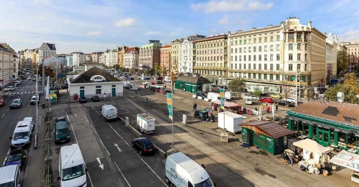 Mehrere Autos parken neben der Straße Menschen spazieren über einen offenen Platz umgeben von gründerzeitlichen Gebäuden und Marktständen helle Fassade mit Bäumen und städtischer Umgebung im Hintergrund
