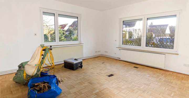 Werkzeuge und Materialien für Bodenarbeiten auf einem Parkettboden in einem leeren Raum mit weißen Wänden zwei großen Fenstern und Heizkörpern im Hintergrund.