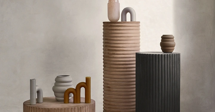 Keramische Vasen und Skulpturen stehen auf drei zylindrischen Säulen in verschiedenen Höhen in einem minimalistischen Raum mit Betonboden und grauen Wänden.