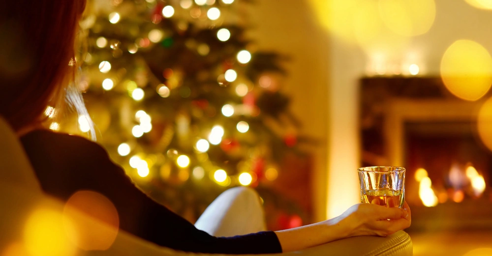 Person sitzt auf einem Sessel, hält ein Glas Getränk, vor einem bunt geschmückten Weihnachtsbaum und einem Kaminfeuer im Hintergrund in gemütlicher Wohnzimmeratmosphäre.