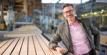 Ein Mann lehnt lächelnd gegen einen Stapel von Holzbrettern in einer großen Werkhalle mit Maschinen im Hintergrund.