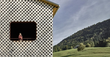 Eine Frau steht auf einem Balkon eines Gebäudes mit einer hölzernen Gitterfassade in einer ländlichen Gegend mit einer grünen Wiese und bewaldeten Hügeln im Hintergrund.