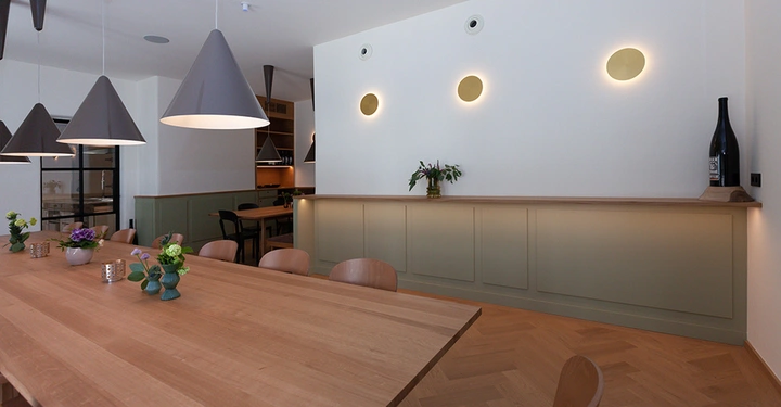 Galerie-Bild 9: Holztisch mit Blumenarrangements steht in modernem Raum mit Hängependelleuchten und Wandverkleidungen beleuchtet durch runde Wandlampen drei. Ein Regal mit Dekor im Hintergrund offene Küche auf der linken Seite.