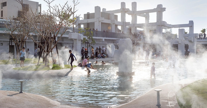 Galerie-Bild 3: Kinder spielen im flachen Wasser eines Parks umgeben von künstlichen Nebelsprühgeräten und kahlen Bäumen vor einem halbfertigen Betongebäude. Mehrere Menschen beobachten von den Seiten.