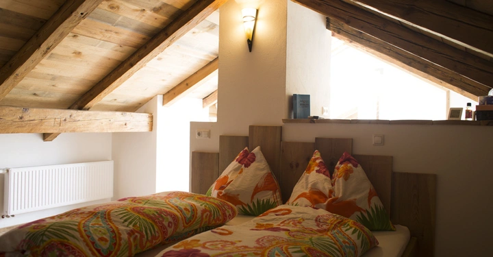 Galerie-Bild 6: Doppelbett mit bunter Bettwäsche ruht unter einem schräge Holzdach in einem rustikalen Schlafzimmer. Ein Wandlicht beleuchtet den Raum. Im Hintergrund sind ein Fenster und Heizkörper sichtbar.