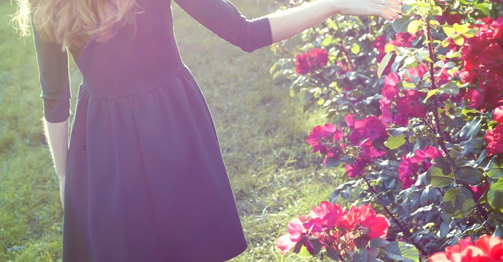 Eine Person mit langem blondem Haar trägt ein grünes Kleid und streicht mit der Hand über rote Rosen in einem sonnigen Garten.