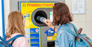 Zwei junge Frauen legen eine Mineralwasserflasche aus transparentem Plastik in einen gelb-blauen Pfandautomaten.
