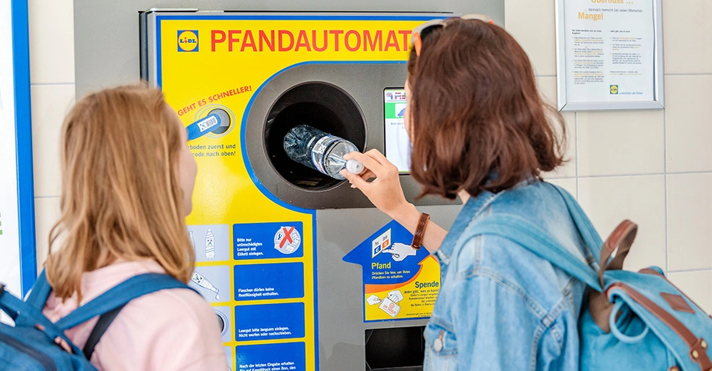 Zwei junge Frauen legen eine Mineralwasserflasche aus transparentem Plastik in einen gelb-blauen Pfandautomaten.
