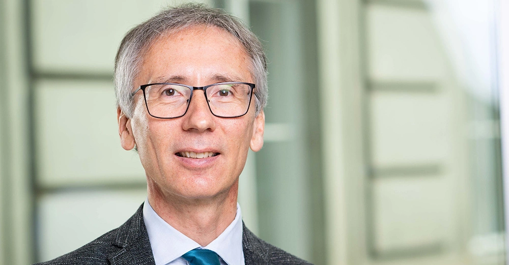 Ein Mann mit Brille und grauem Haar lächelt leicht im Vordergrund eines Büros mit verschwommener heller Inneneinrichtung im Hintergrund.