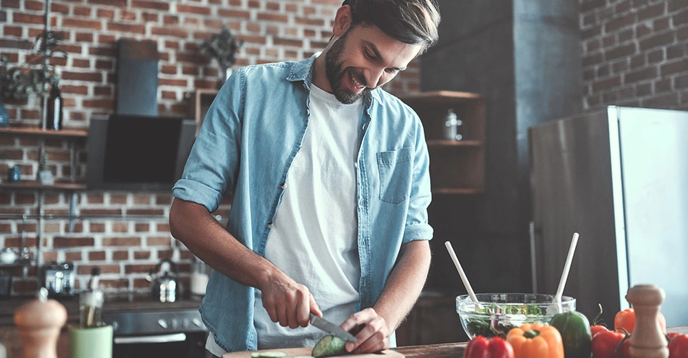 Junger Mann in weißem Shirt und blauem offenen Hemd schneidet in der Küche auf Holzbrett lächelnd Gurken. 