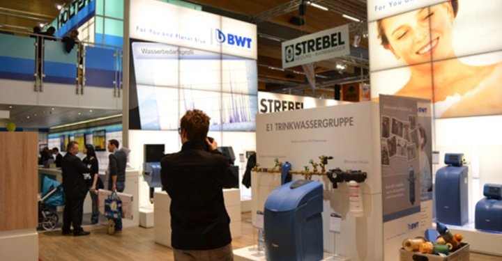 Galerie-Bild 19: Person macht ein Foto einer blauen Wasserfilteranlage auf einer Messe mit mehreren Ständen und Displays Etiketten sichtbar: BWT Wasseraufbereitung STREBEL E1 TRINKWASSERGRUPPE For You and Planet Blue