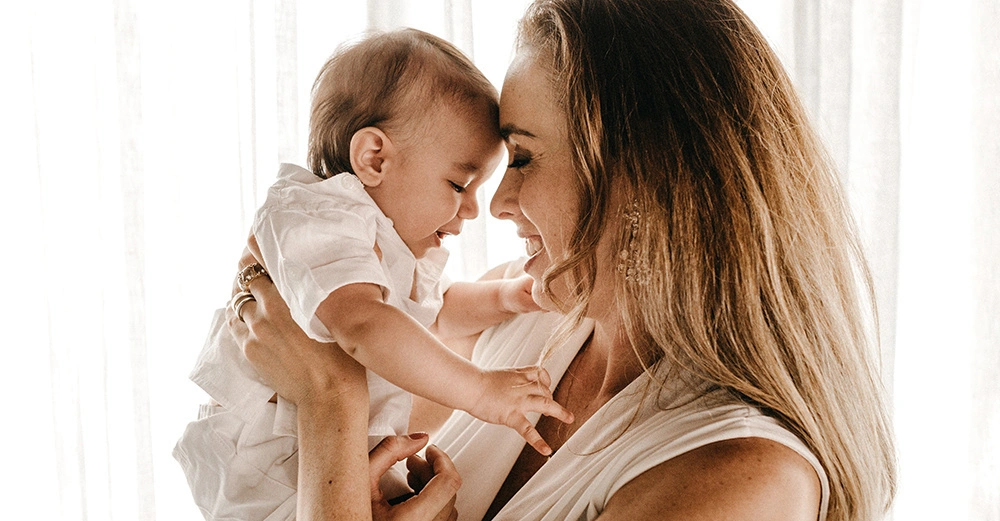 Eine Frau hält ein Baby im Arm und beide lächeln sich an; sie stehen vor einem hellen Vorhang in einem gut beleuchteten Raum.